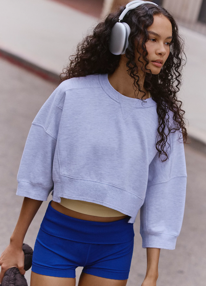 A woman wearing a light heather grey crewneck sweatshirt and bright blue athletic shorts, with white over-ear headphones, photographed outdoors against a blurred concrete background.