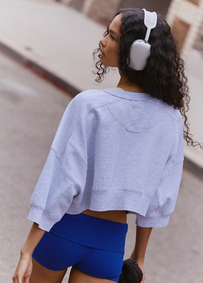 A woman with curly dark hair wearing a heather grey cropped sweatshirt and bright blue athletic shorts, with white over-ear headphones, photographed outdoors on a blurred street background.
