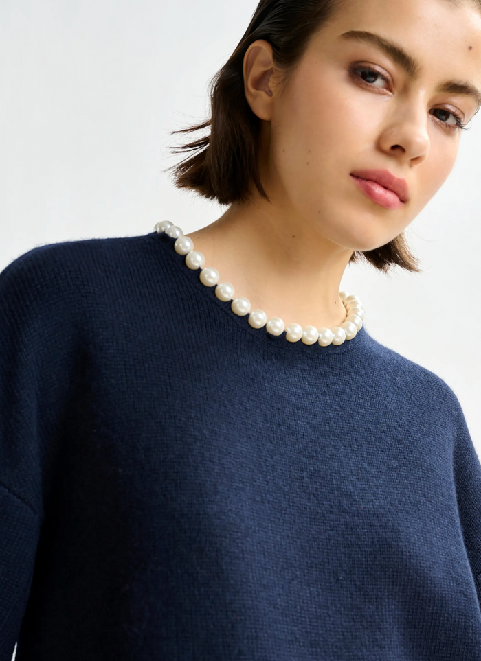 Woman wearing a navy blue sweater and pearl necklace against a white background