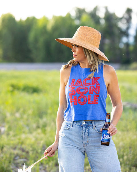 A person wearing a Jackson Hole letter tank top in a field. The tank top is blue with the text 'JACKSON HOLE' in bold red letters.