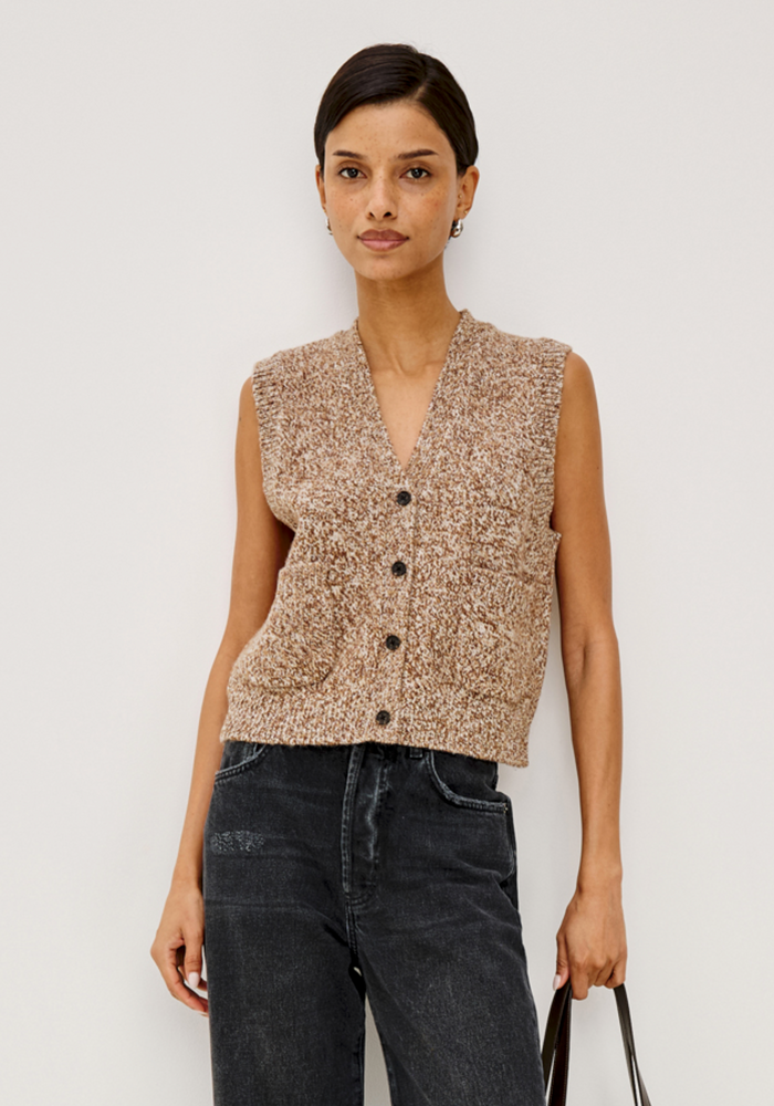 A model wearing a sleeveless, textured knit vest in a beige and brown speckled pattern with black buttons, paired with dark jeans, against a plain light gray background.