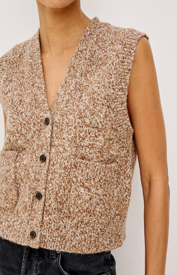 A close-up of a person wearing a sleeveless knit vest with a marled beige and brown texture, dark buttons, and a V-neckline, paired with dark jeans against a plain light background.