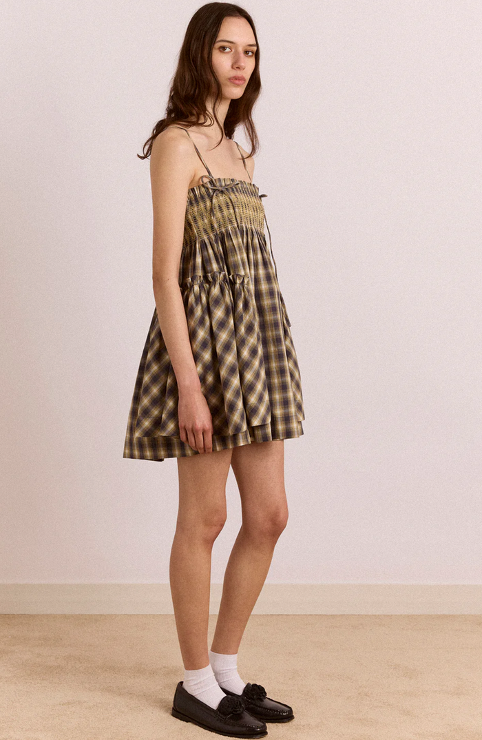 A woman modeling a short plaid mini dress with thin shoulder straps, a smocked bodice, and a gathered flared skirt. She is wearing white socks and black loafers against a plain beige wall and floor.