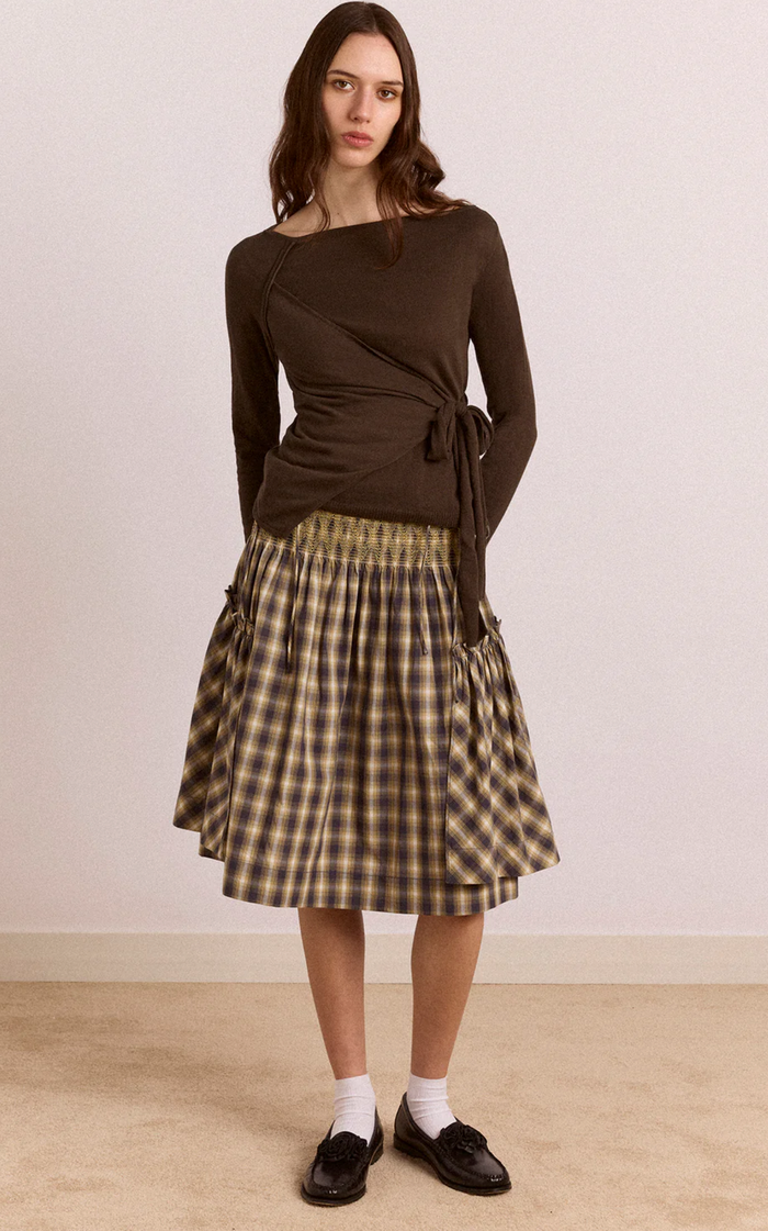 A woman modeling a dark brown asymmetrical long-sleeve wrap top paired with a brown and beige plaid pleated skirt, white socks, and black loafers, standing against a plain off-white wall.