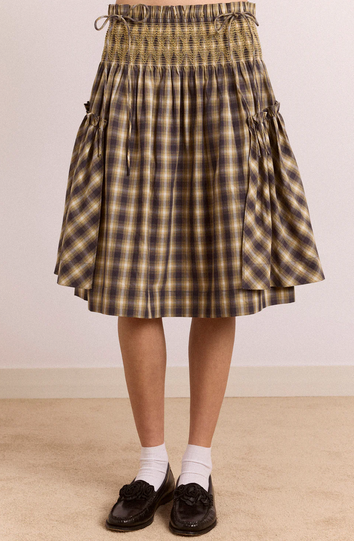 A pleated A-line skirt with a brown, beige, and black plaid pattern, featuring a smocked waistband and side tie details, shown on a model wearing white socks and black loafers.