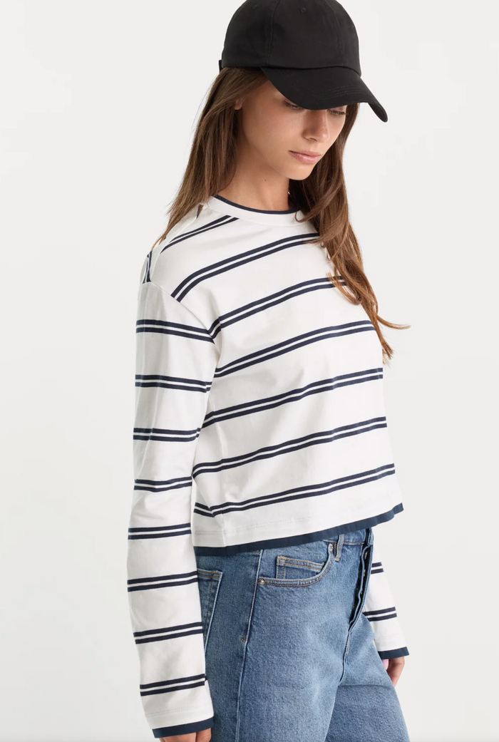 A woman wearing a white long-sleeve cropped top with navy blue horizontal stripes, paired with blue jeans and a black baseball cap, photographed against a plain white studio background.