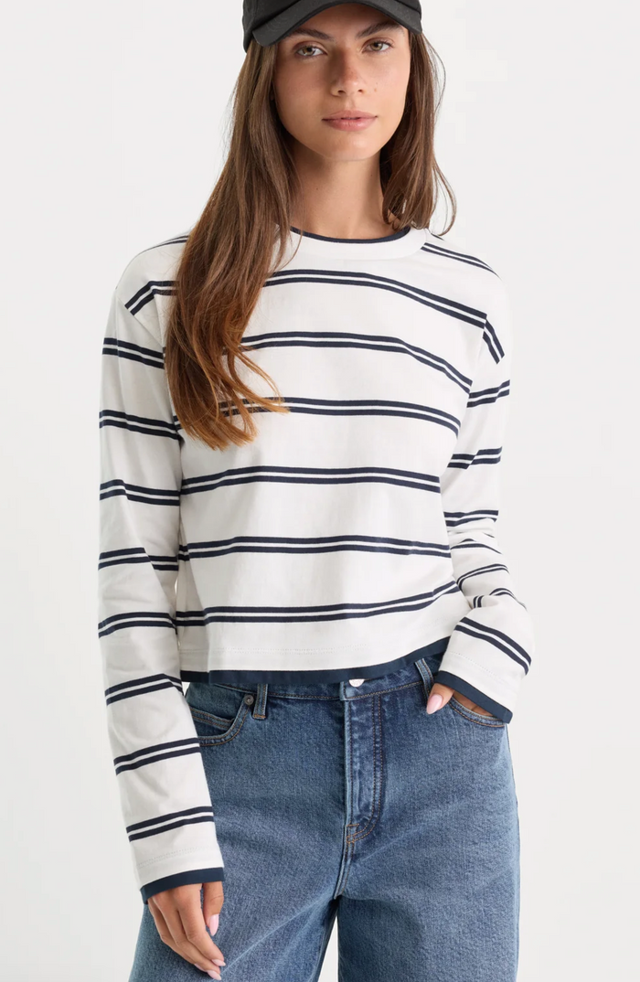 A woman wearing a white long-sleeve crewneck sweater with thin navy horizontal stripes, paired with blue jeans and a black cap, against a plain white studio background.