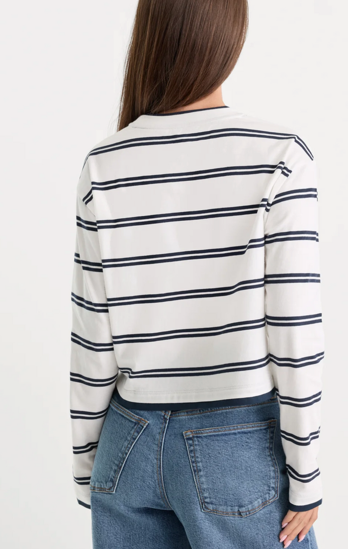 A woman shown from the back wearing a cropped long-sleeve top with horizontal navy blue stripes on an off-white background, paired with blue jeans, against a plain white studio background.
