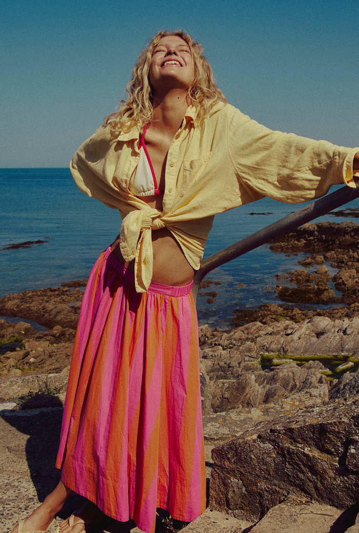 Woman in a yellow shirt and pink striped skirt standing on rocks by the ocean