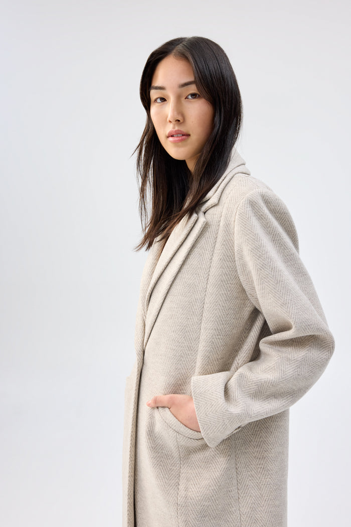 Side profile of a model wearing a beige textured faux wool coat with notch lapel, hand in pocket