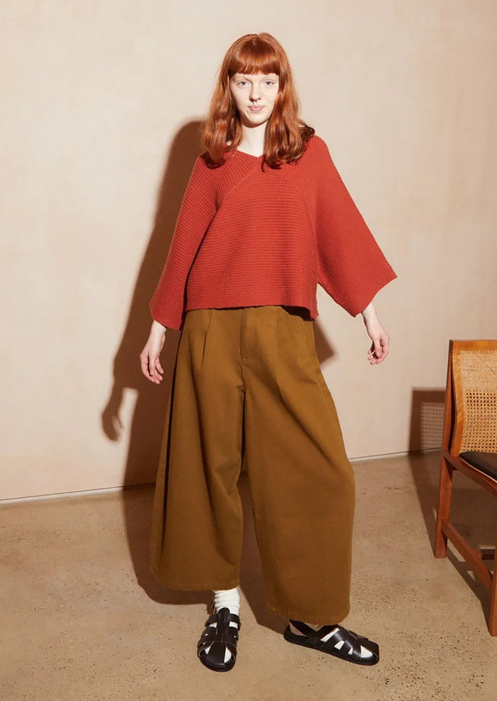 A model with red hair stands in a studio wearing a rust-orange ribbed knit poncho-style top and wide-leg brown trousers, paired with white socks and black sandals. The background is a warm beige wall and floor with a wooden chair partially visible on the right.