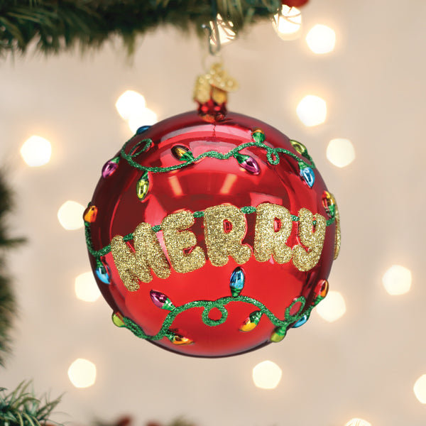 A shiny red Christmas ornament decorated with glittery gold letters spelling MERRY and colorful painted holiday lights, hanging on a tree with blurred white lights in the background.