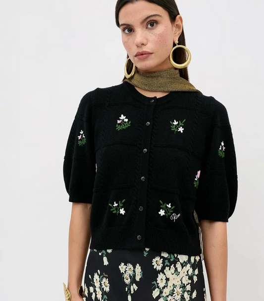 A model wears a black short-sleeve knit cardigan featuring embroidered white flowers and green leaves on the front and pockets, styled with a textured olive green scarf and a black floral skirt against a plain white background.