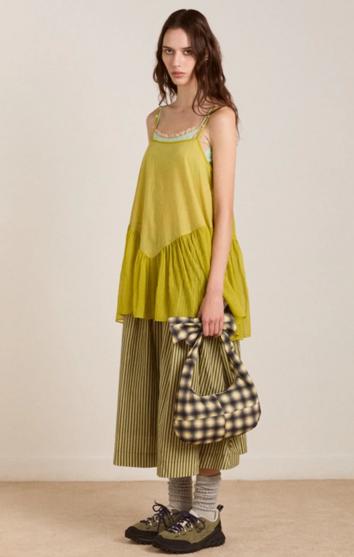 A model stands against a plain beige studio wall wearing a loose chartreuse sleeveless top, striped green and beige wide-leg pants, striped socks, and hiking shoes, while holding a large black-and-beige gingham tote bag.
