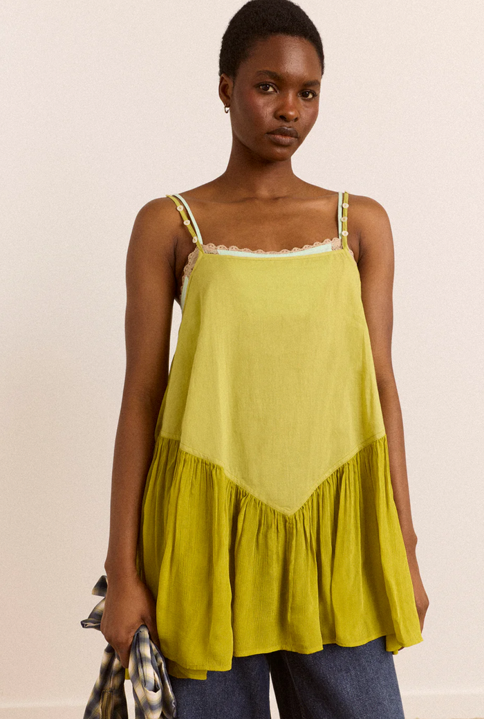 A woman modeling a chartreuse tiered camisole top with thin straps and a ruffled hem, paired with blue jeans against a plain off-white background.