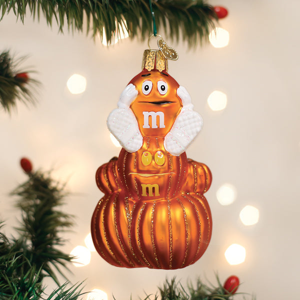 An orange M&M ornament with big white gloves and a surprised expression hangs on a Christmas tree, surrounded by warm, blurred holiday lights and green pine branches.