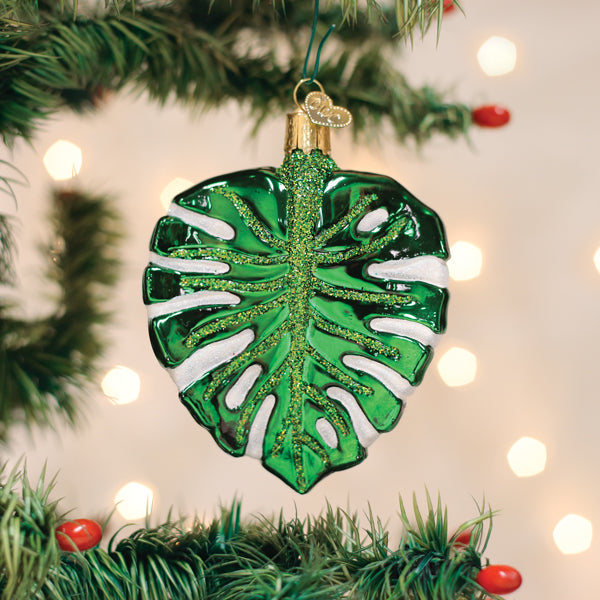 A shiny glass ornament shaped like a green monstera leaf with white accents hangs on a Christmas tree branch, surrounded by twinkling lights and red berries.