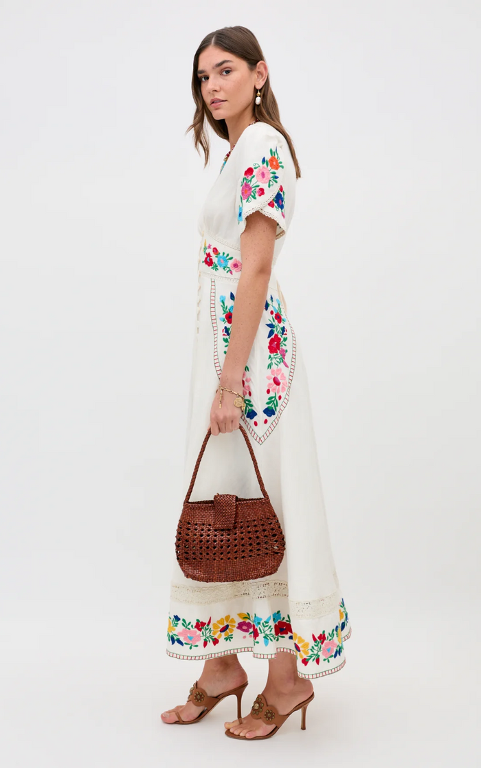 Woman wearing a white dress with colorful floral embroidery, holding a brown handbag on a white background
