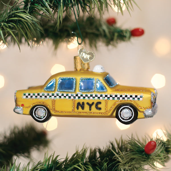 A yellow NYC taxi cab ornament with black checkered details hangs on a Christmas tree, surrounded by green pine needles and red berries, with blurred festive lights in the background.