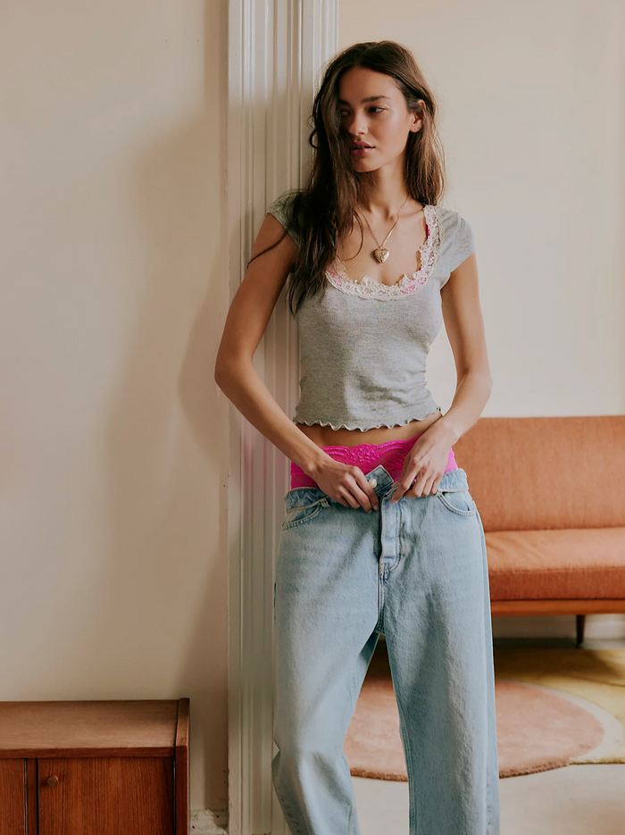 Woman standing indoors wearing a light gray crop top and blue jeans, leaning against a wall.