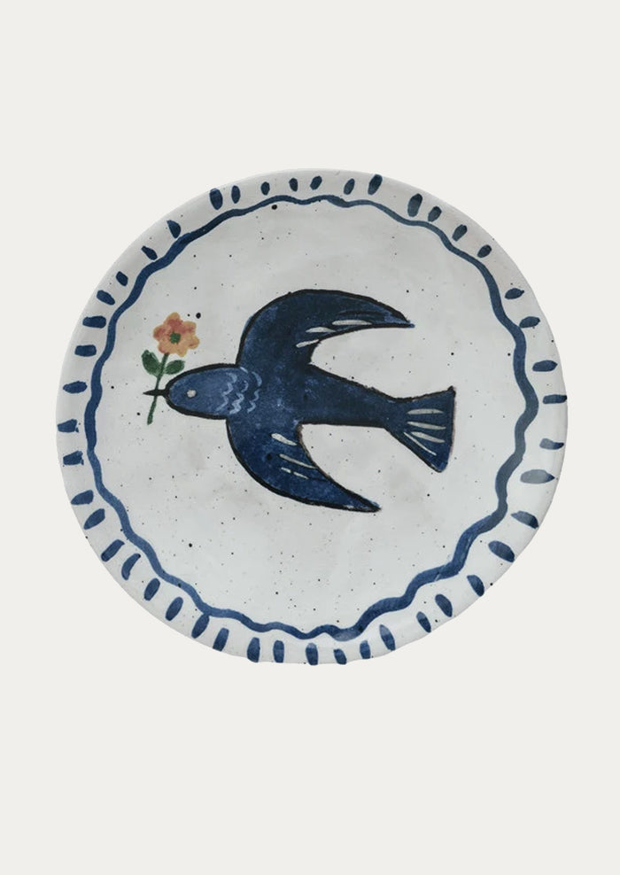 A small ceramic plate with hand painted imagery of blue bird holding flower.