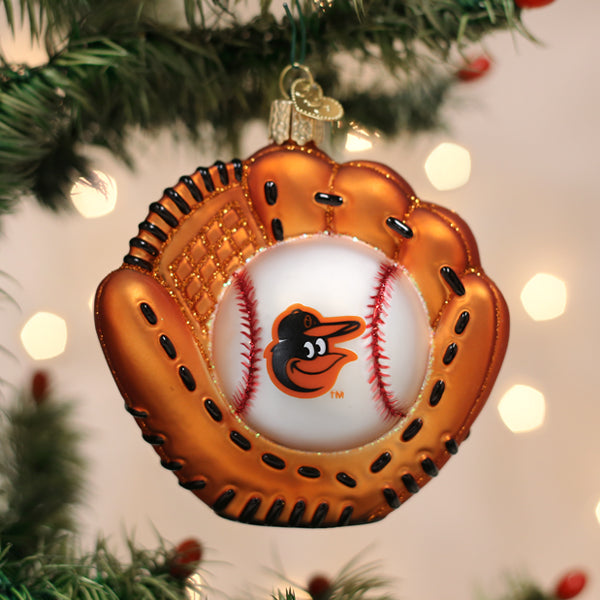 A Christmas tree ornament shaped like a baseball glove holding a baseball with the Baltimore Orioles logo, hanging on a green tree with blurred lights in the background.