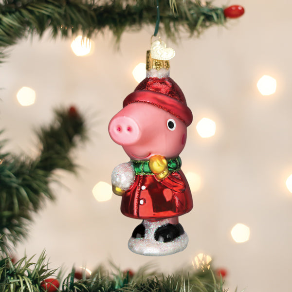 A Christmas tree ornament shaped like Peppa Pig wearing a red festive coat and hat, holding a snowball, hanging on a decorated tree branch with blurred lights in the background.