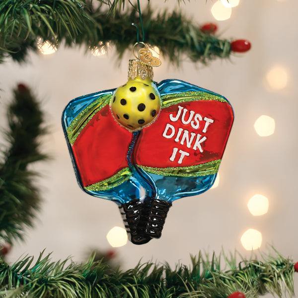A Christmas tree ornament shaped like two pickleball paddles with the words JUST DINK IT and a yellow pickleball hanging above, surrounded by greenery and holiday lights in the background.