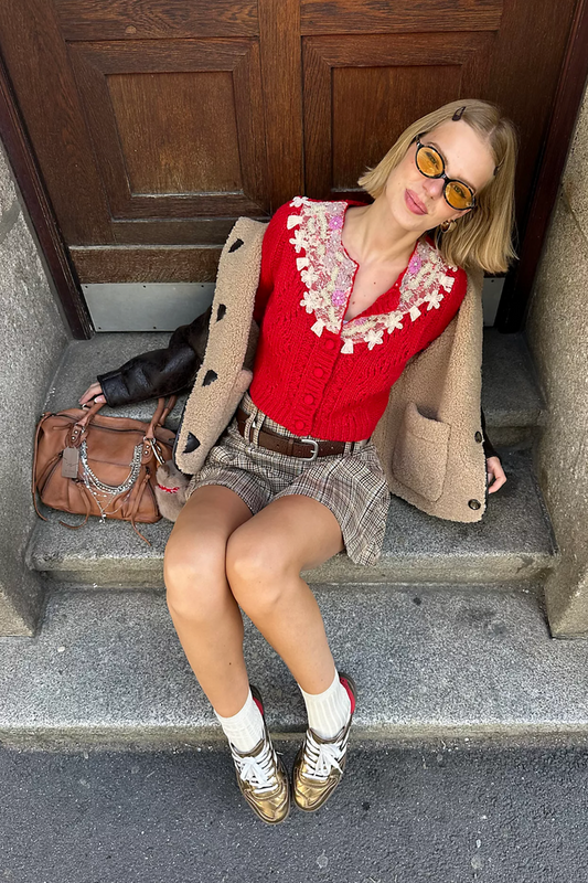 Woman sitting on steps wearing a red cardigan sweater and beige coat, with a brown handbag beside her.