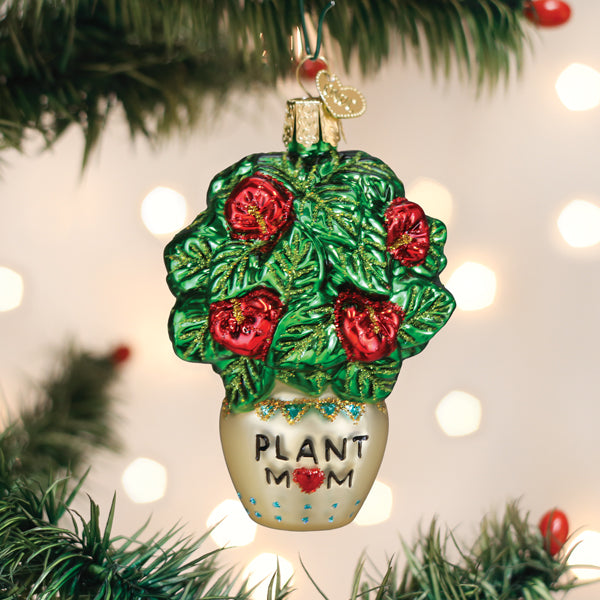 A glass Christmas tree ornament shaped like a potted plant with green leaves and red flowers. The pot has PLANT MOM written on it with a red heart, and is hanging on a Christmas tree branch.