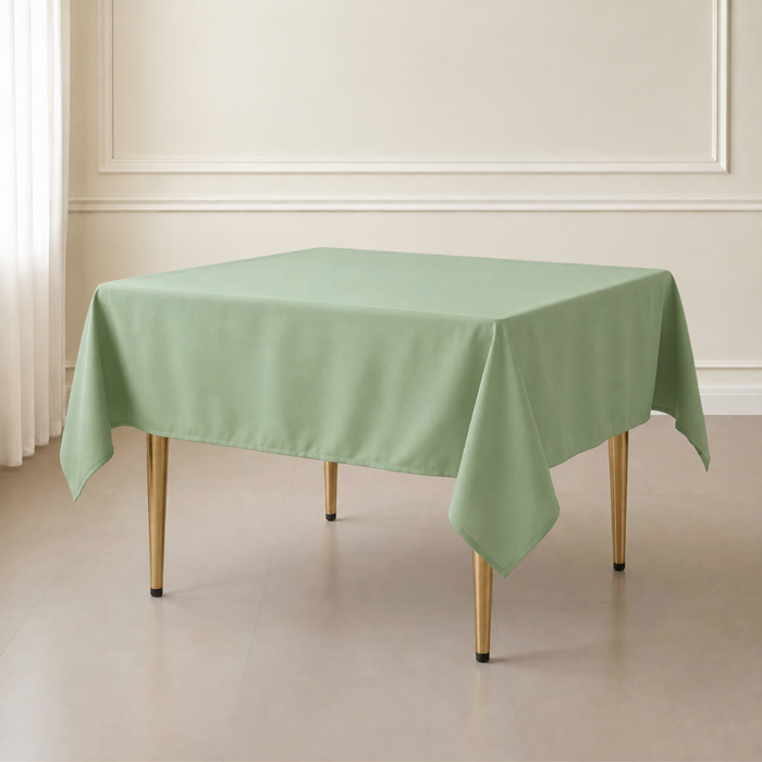 A sage green rectangular tablecloth draped over a table with gold-toned legs, set in a minimalist room with cream walls and a light floor.