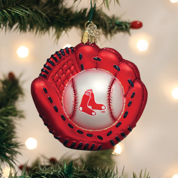 A festive ornament shaped like a red baseball glove holding a baseball with the Boston Red Sox logo hangs on a Christmas tree with blurred white lights in the background.