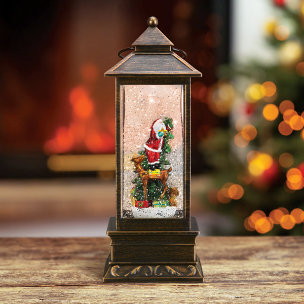 A bronze lantern-shaped snow globe with a Santa Claus figurine inside stands on a wooden surface. In the background, there are blurred Christmas lights and a lit fireplace.