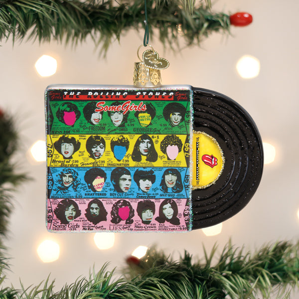 A Christmas tree ornament shaped like a colorful vinyl record and album cover, featuring illustrated faces and the text Some Girls. The ornament hangs on a tree with green branches and white lights in the background.