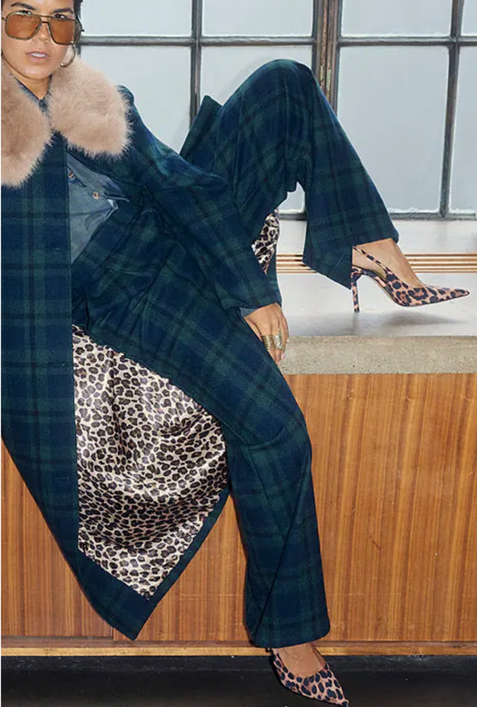 Woman in a blue plaid coat with fur collar and leopard print pants sitting on a wooden bench.