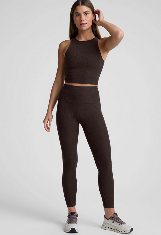 A woman modeling a matching chocolate-colored activewear set consisting of a sleeveless ribbed crop top and high-waisted leggings, paired with gray and white sneakers, against a light gray studio background.