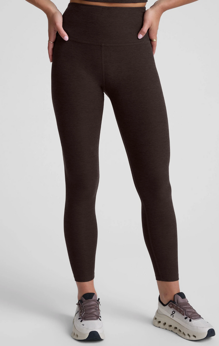 A person wearing chocolate-colored high-waisted leggings with side pockets, paired with white and gray sneakers, shown from the waist down against a light gray studio background.