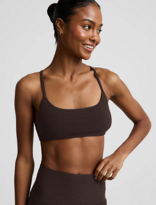 A woman wearing a chocolate-colored scoop-neck athletic sports bra and matching high-waisted bottoms, photographed against a solid light gray studio background.