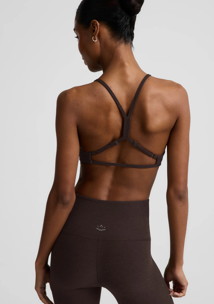 Rear view of a woman wearing a chocolate-colored sports bra with crisscross back straps and matching high-waisted leggings against a light gray studio background.