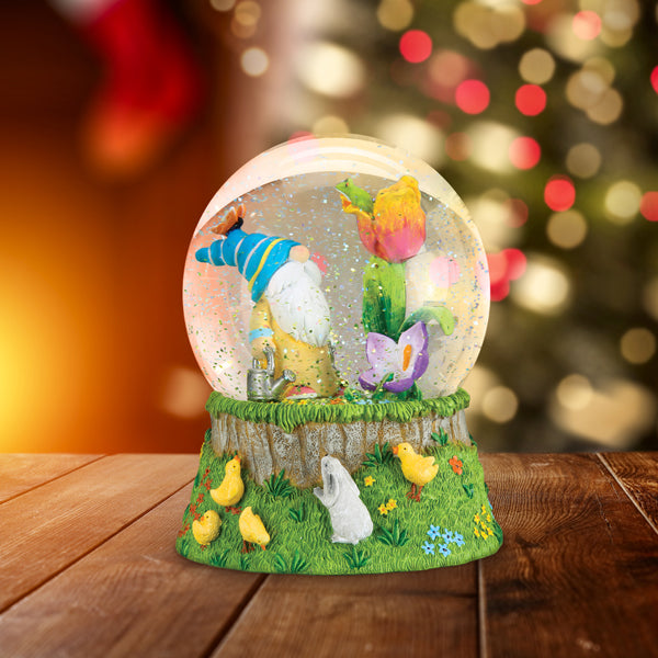 A whimsical snow globe with a gnome in a blue hat, colorful flowers, and yellow chicks sits on a wooden surface, with warm, blurred holiday lights in the background.