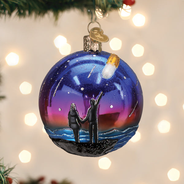 A Christmas tree ornament painted with a sunset sky, stars, and comet. Silhouettes of two people, one pointing upward, stand together on a hill, with blurry holiday lights in the background.