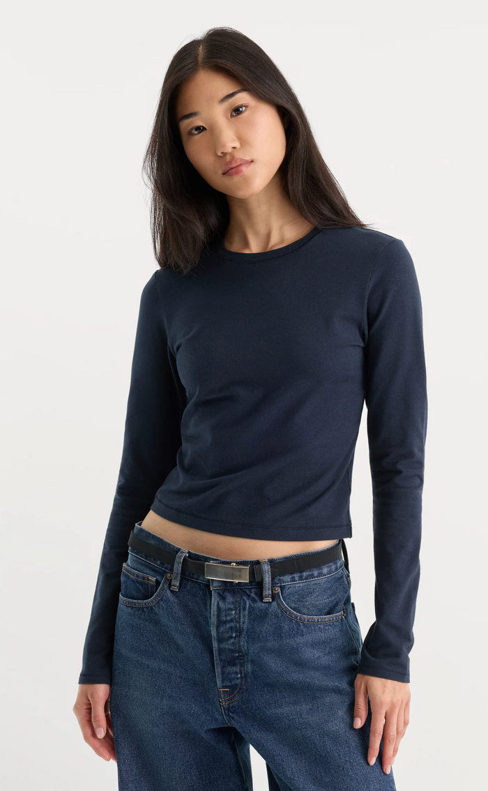 A woman modeling a dark navy blue long-sleeve crop top with a crew neck, paired with blue jeans, against a plain light gray background.