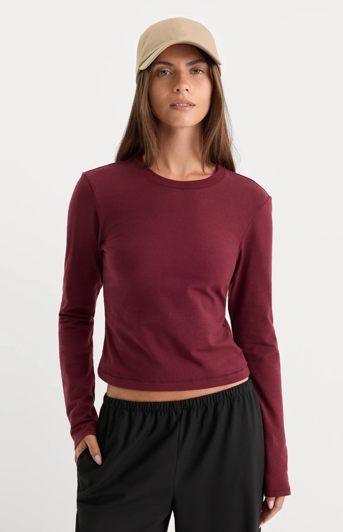 A woman modeling a long-sleeve crewneck crop top in burgundy, paired with black sweatpants and a beige baseball cap, against a plain light gray background.