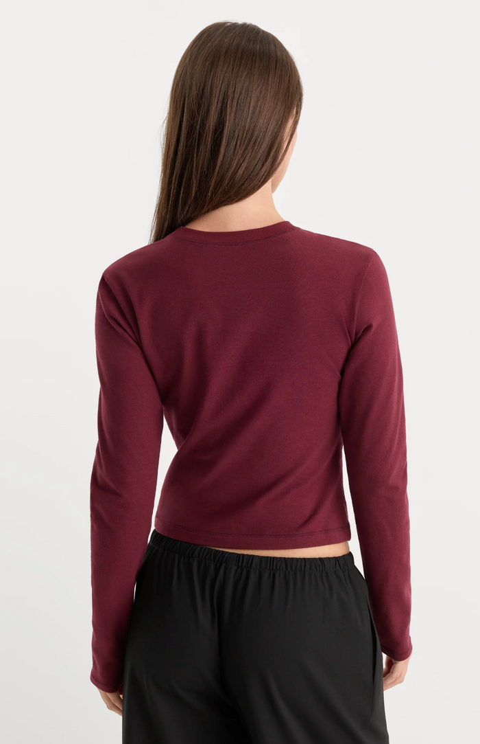 A woman viewed from the back wearing a long-sleeve cropped top in burgundy and black pants, against a plain light gray studio background.