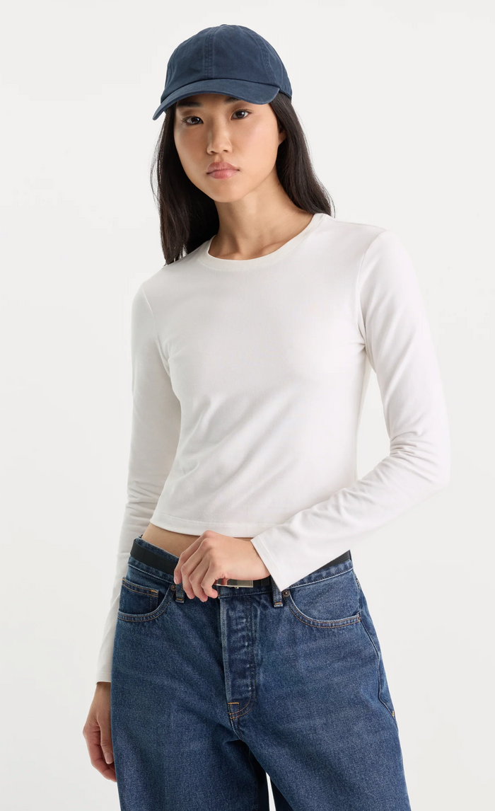 A woman wearing a fitted white long-sleeve crewneck top, blue jeans, and a navy blue baseball cap, photographed against a clean white studio background.