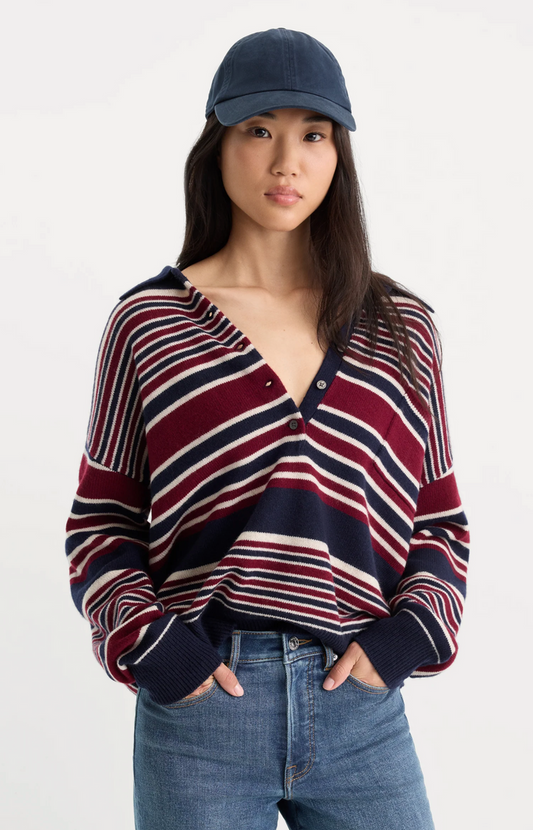 A woman wearing a navy, burgundy, and cream striped knit sweater with a V-neck and button detail, paired with blue jeans and a navy baseball cap, against a plain light gray studio background.