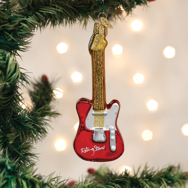 A red and gold electric guitar ornament with Rolling Stones written on it hangs on a green Christmas tree branch, with soft white holiday lights in the background.