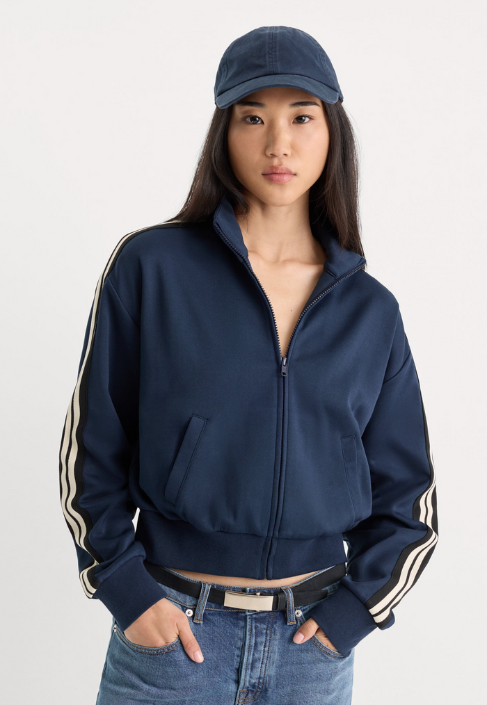 A woman wearing a navy blue cropped zip-up track jacket with cream stripes down the sleeves, paired with a matching navy baseball cap and blue jeans, photographed against a light gray studio background.