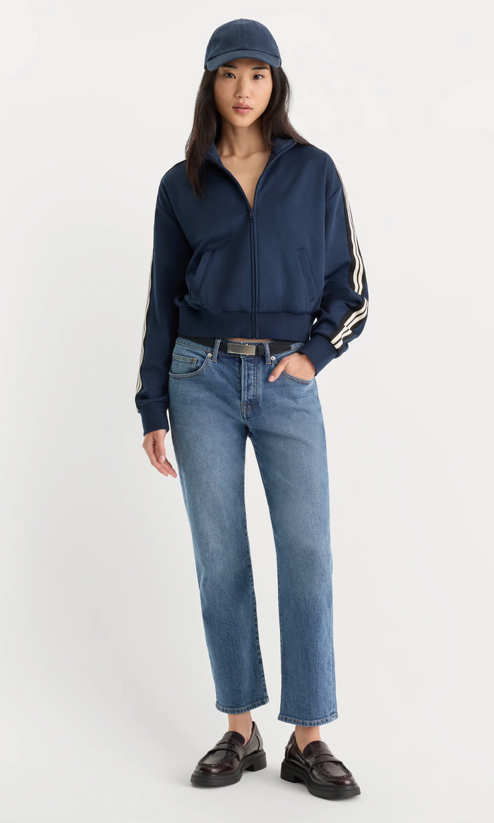 A model wearing a navy blue zip-up track jacket with white stripes on the sleeves, blue jeans, a navy cap, and black loafers, standing against a light gray studio background.