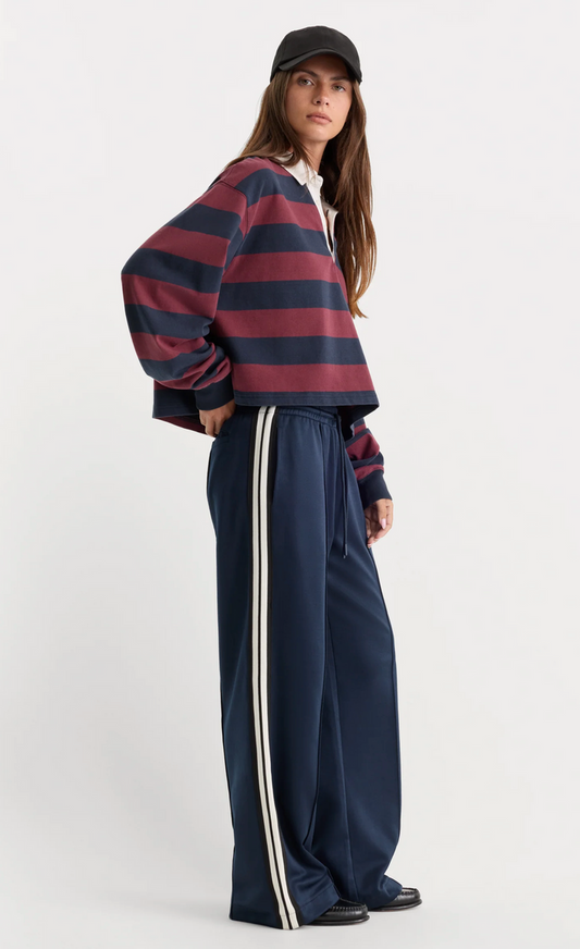 A model wearing a cropped, long-sleeve striped knit top in navy and burgundy, paired with navy wide-leg track pants featuring white side stripes, a black baseball cap, and black shoes, against a plain light gray studio background.